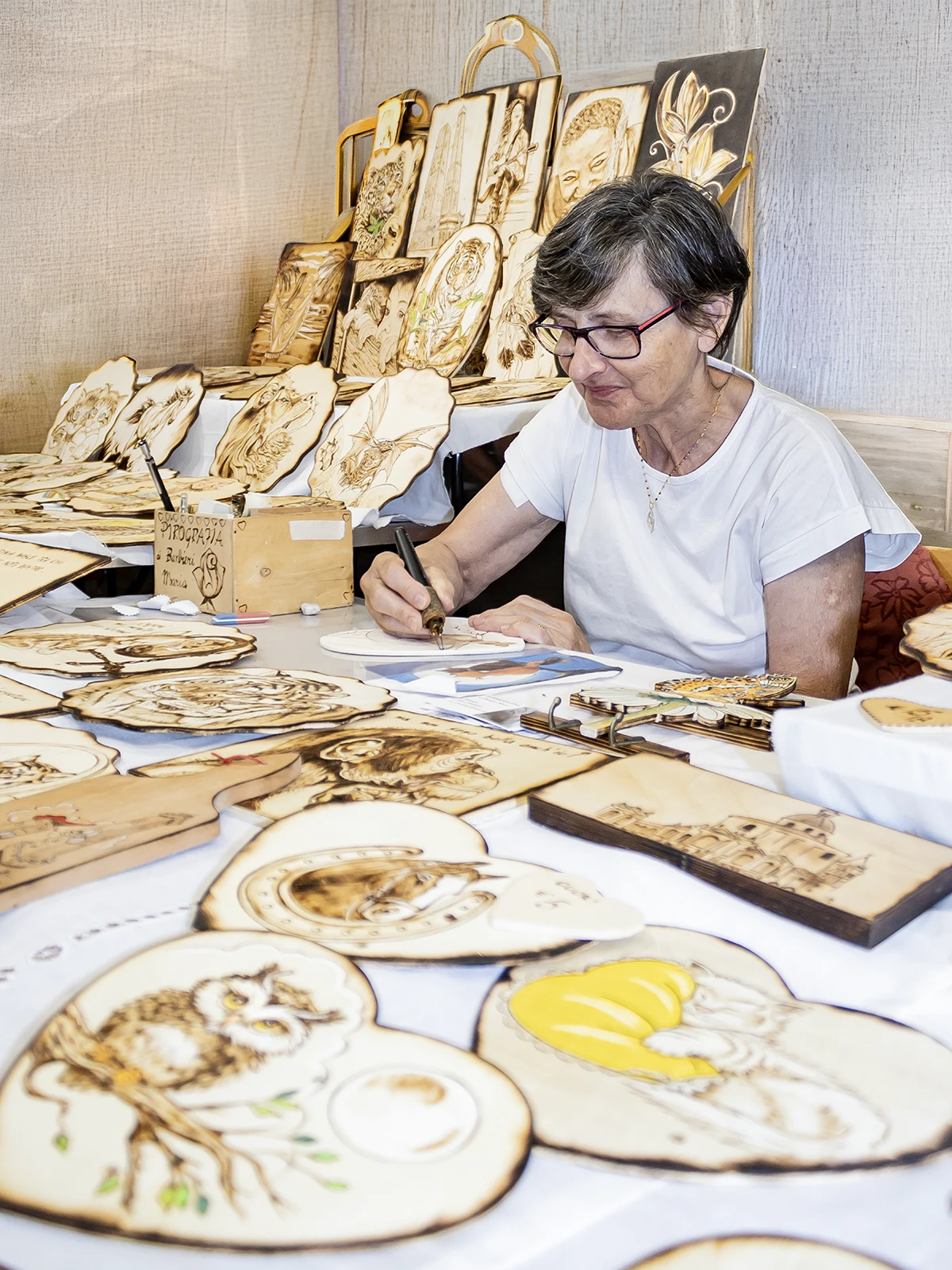 Maria Barbieri, pirografa, al lavoro con la penna termica su una tavoletta di legno tra le sue opere esposte