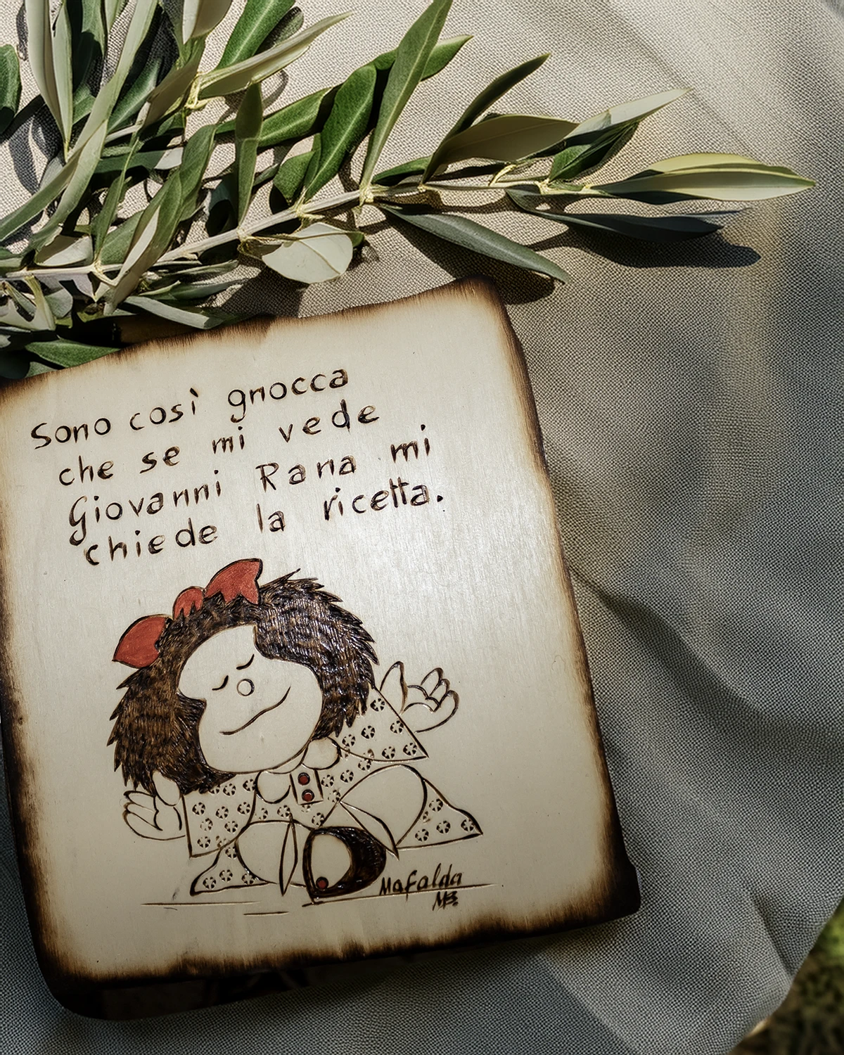 Tavoletta pirografata con Mafalda e una frase ironica, posata su un panno chiaro con un ramo d’ulivo